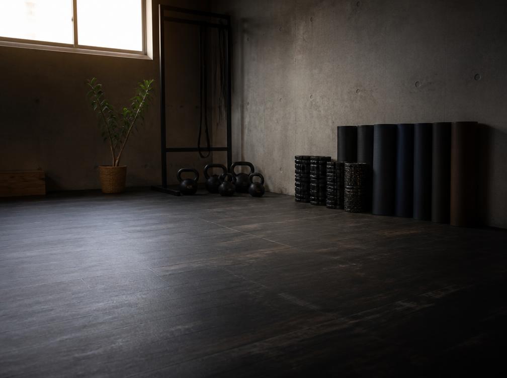 Rolled mats, foam rollers and kettlebells stacked against a concrete wall on an open studio floor